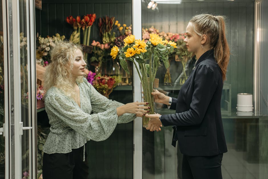 A woman with curly blonde hair, dressed in a light-colored floral blouse, hands a bouquet of yellow sunflowers wrapped in clear plastic to a female florist with straight, light brown hair tied in a po