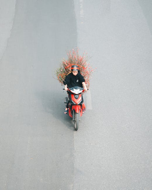 A delivery person riding a red scooter on an urban street during daytime, with a large bouquet of flowers secured on the back of the scooter. The bouquet features various flowers in shades of pink, re