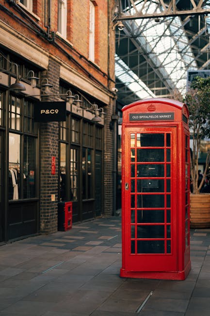 The image depicts a vintage red British telephone booth situated outdoors near a brick building with large glass windows, part of Old Spitalfields Market. The booth's vibrant color contrasts against t